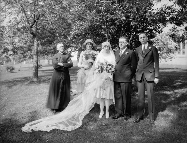 Don Kennedy & Eleanor Rusch and their wedding attendants poing with the priest outdoors.
