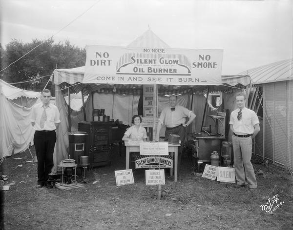 Sales personnel pose in the Silent Glow oil burner booth with  an oil burning kitchen stove, furnaces, and portable styles of oil burners at the Dane County Fair.