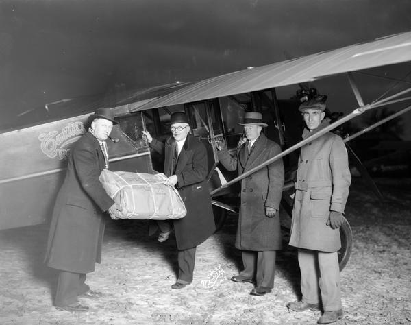 Four men are standing beside a Curtiss airplane to receive a parcel flown in from the National Theatre Supply Company for the Eastwood Theatre.