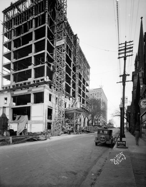 View looking up Fairchild Street towards the Wisconsin Power and Light building, at 122 W. Washington Avenue, under construction, with a man working near a materials elevator and mixer. On the right is the First Congregational Church, and the Loraine Hotel is up the hill in the background across West Washington Avenue.