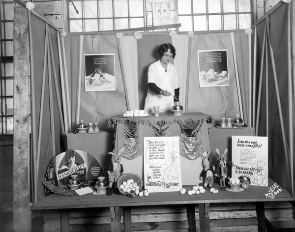 A woman demonstrates how to use a Hankscraft egg cooker in a  mock Easter display window with Easter bunnies and Easter eggs.