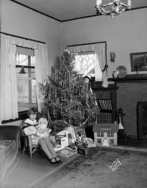 A little girl posing in a living room with a Christmas tree, toys, and a doll she got for Christmas.