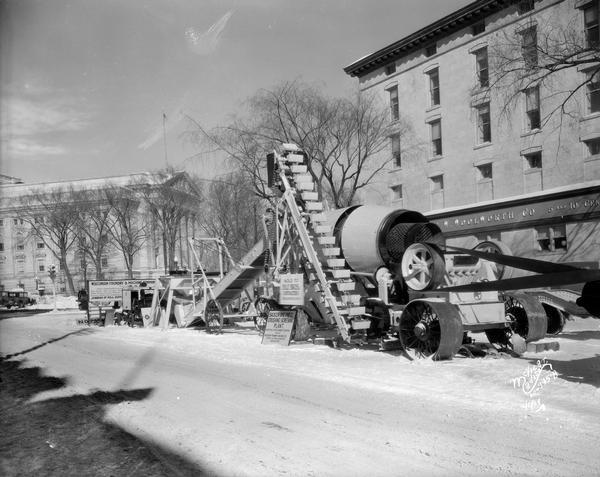 A Wisconsin Foundry & Machine Company road equipment display of a Badger one-piece crushing screening plant. The equipment is displayed on Monona Avenue. Snow is in the ground. The Woolworth's building and the Wisconsin State Capitol are in background.