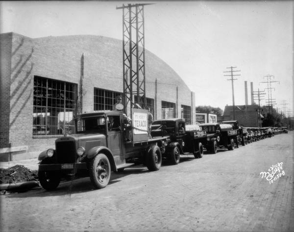 View down street towards 23 trucks and cars in a parade celebrating the completion of the take over of the Capitol Oil Company by Texaco, located at 919 East Main Street.