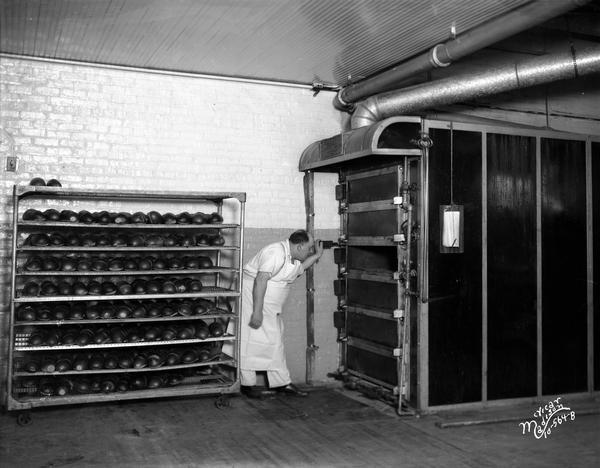 A baker is looking into the oven at Gardner Baking Company, with a rack of loaves of bread cooling beside him.