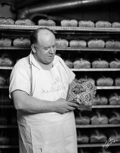Baker holding a loaf of Gardner's Double Duty bread with shelves of bread in the background. The baker is wearing Else Russell's Best Flour apron.