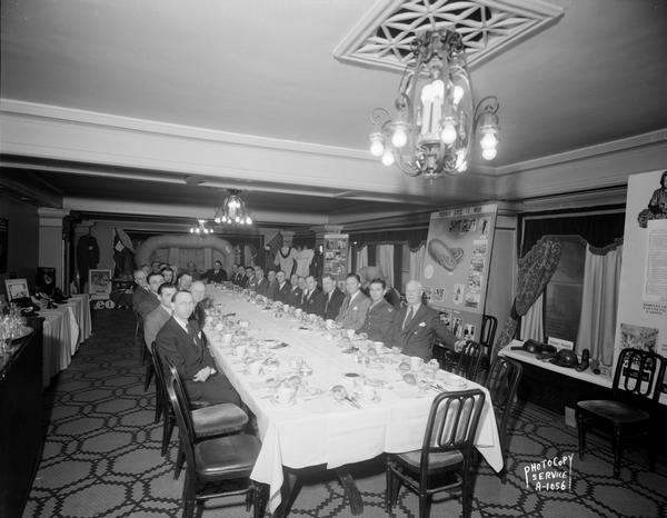 United States Rubber Company group seated at a table with a display of rubber products in the background, "Rubber Goes to War," in the Pompeii Room at the Loraine Hotel, 119-125 West Washington Avenue.