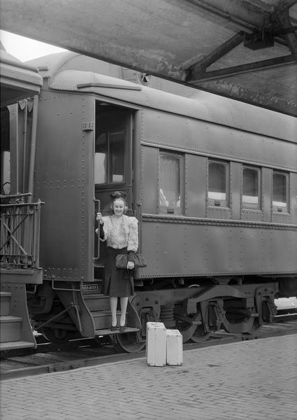 Manchester's Department Store coed model, wearing a fur jacket, hat and purse, posing on the steps of a passenger train, with two pieces of luggage on the ground.