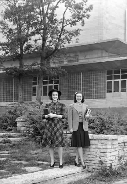 Two Manchester's Department Store coed models, carrying books, one wearing a dress and hat, the other a jacket, skirt and sweater, pose beside the Memorial Union on the University of Wisconsin, Madison campus.