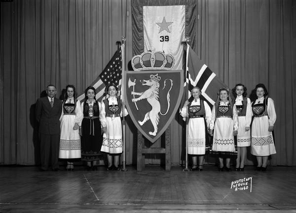 Norwegian-American Society members in costume on stage at Central High School with United States and Norwegian flags and Norwegian coat of arms.