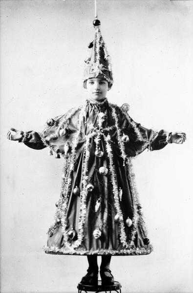 Girl standing on stool. She is wearing a dress and hat with tinsel and glass ornaments.