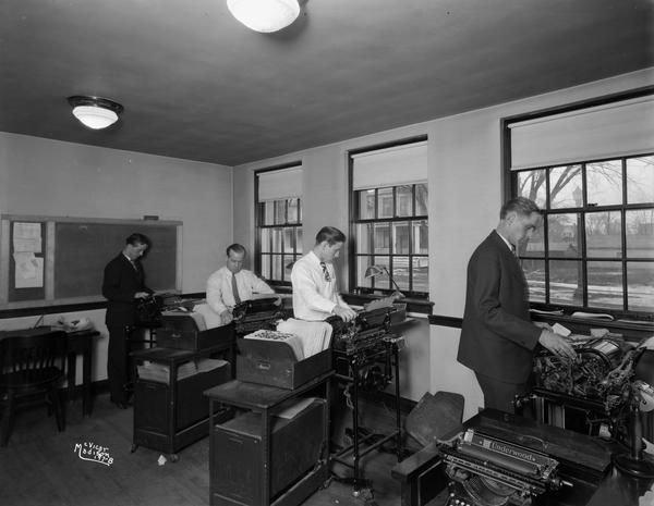 Four men in the bookkeeping department at the Security State Bank, 1965 Atwood Avenue. They are standing and working at machines near a row of windows that look out into the street.
