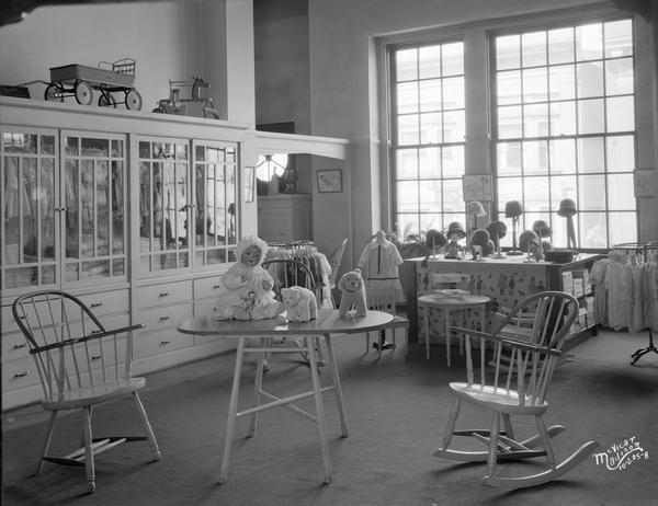 Kessenich's infant department. In the foreground is a table with a doll and stuffed toys on it, surrounded by three rocking chairs. Along the wall in the background is a large, glass cabinet with clothes on display inside, and on top of the cabinet is a toy wagon and a spring rocking horse. Hats are on a large table in front of the windows in the background.