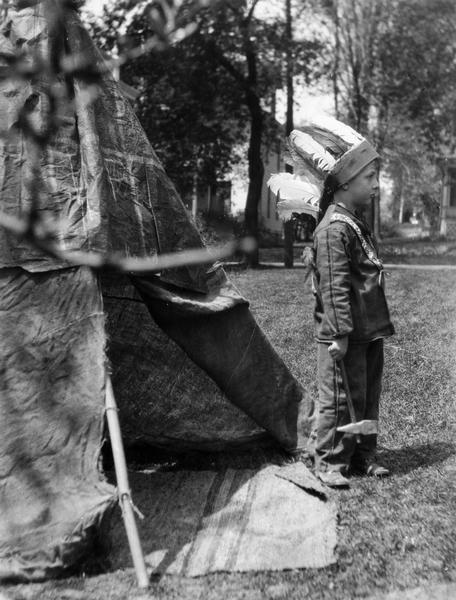A boy is standing at the entrance to a teepee. He is dressed in Native American clothing, and is holding an axe.