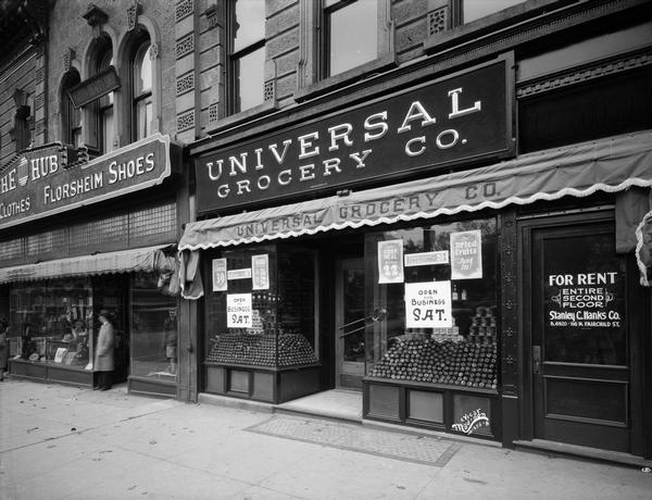 Storefront of the Universal Grocery Company at 20 W. Mifflin Street, with window displays. On the left is a portion of The Hub, 22 W. Mifflin Street, a store for men's clothes and Florsheim Shoes. On the right is a doorway with a sign that reads: "For Rent, Entire Second Floor, Stanley C. Hanks, Co."