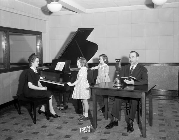 Reverend C.O. and Mrs. Grobber and daughters at WIBU radio station studio. Reverend Grobber is sitting in front of a microphone, Mrs. Grobber and daughters are at the piano.