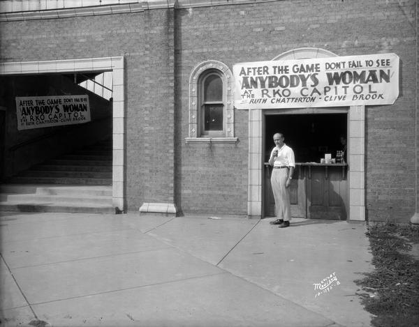 Motion picture advertisement for RKO Capitol Theatre's "Anybody's Woman" displayed at Breese Stevens Field. A man is standing at the concession stand below sign.