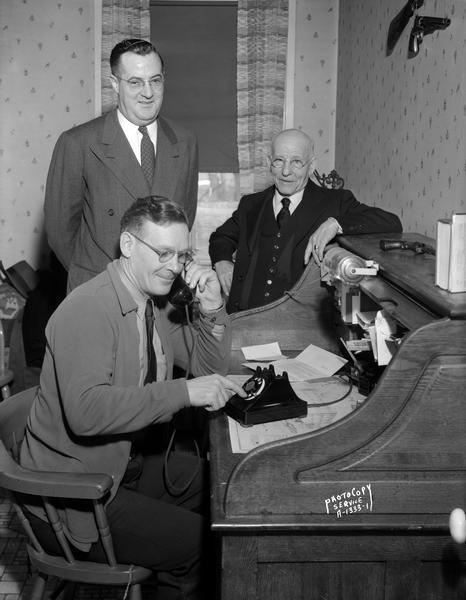 Mazomanie Village President Theodore R. King, seated at a roll top desk, dials the first telephone call to be handled through the Wisconsin Telephone Company's new dial central office in Mazomanie.  Witnessing the historic event are T.J. Murphy, standing at left, Madison district manager for the Wisconsin Telephone Company, and Frank Lashway, prominent resident of Mazomanie who served as the company's agent in Mazomanie for many years before retiring a few years previous to this date. This telephone call was received by telephone company Mazomanie agent Charles T. Davies at Davies & Son Store.