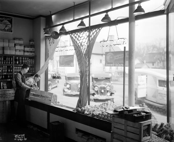 Morning view of the interior of Frank Bros. grocery store, located at 609-613 University Avenue, with two shop keepers stocking the window with Seald Sweet oranges. Seen through the front window are cars and trucks parked along the curb at an angle.