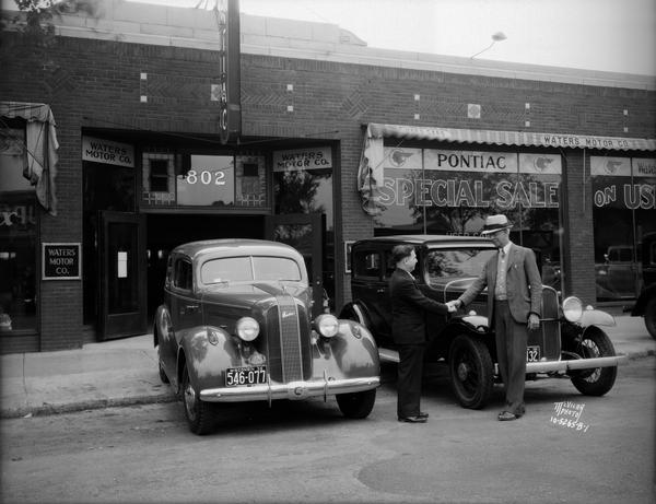 Waters Motor Co., located at 802 East Washington Avenue. There are two men shaking hands in front of old and new Pontiacs.