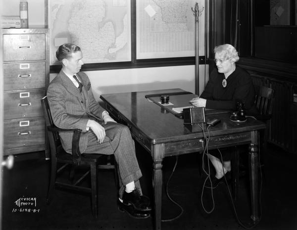 Regina E. Groves (Mrs. Earl Barnhart) talking to a man sitting across the table/desk from her at the Groves School for Secretaries, located at 502 State Street.