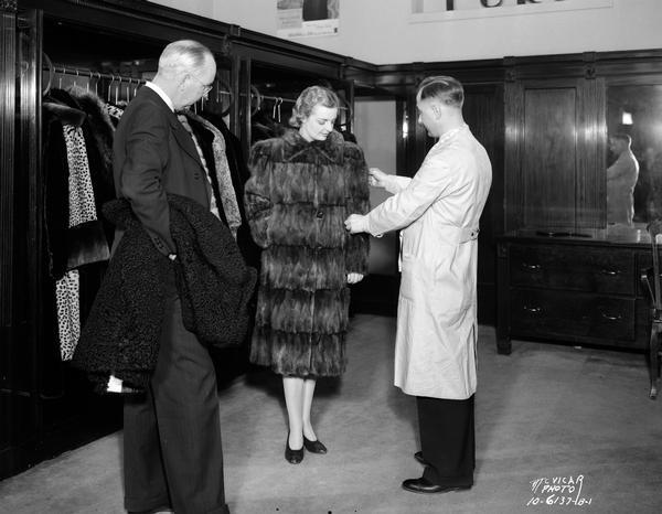 Salesman helping woman trying on fur coat, with her husband looking on, at Simpsons Garment Co., located at 23-25 N. Pinckney Street.
