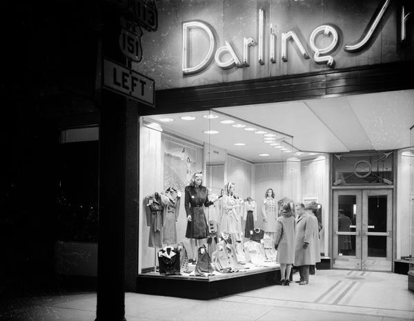Exterior night view of the Darling Shop, located at 9 East Main Street, with people looking at the window displays. A portion of the lighted neon "Darling Shop" sign is visible as is a street lamp with highway signs for U.S. Highway 151 and State Highway 113.