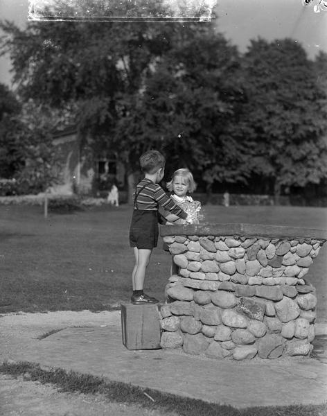 Boy and girl modeling Manchester's Department Store clothes at the Vilas Park drinking fountain. Included in sleeve is a separate negative overlay negative with the words "'A Hundred to One' It's for You!" Advertisement says "One to Six year olds will find their needs at pleasant low prices in the Baby Shop on the Third Floor."