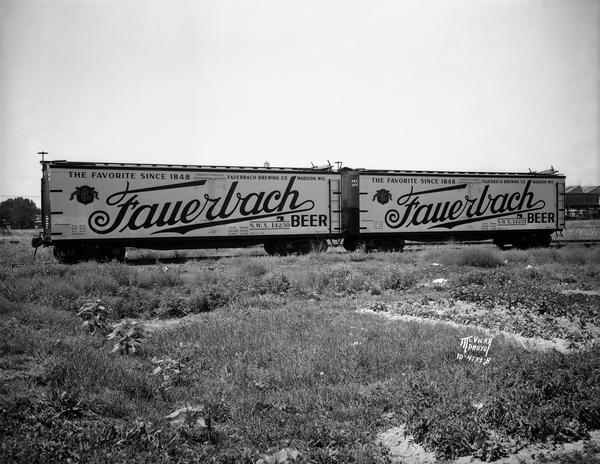 Two railroad freight cars with Fauerbach Beer signs covering their sides are parked outdoors on railroad tracks in a railroad yard.