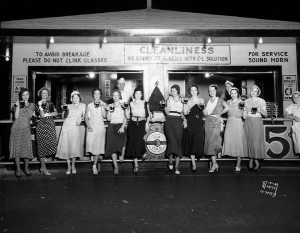 Fanchon and Mario "Rhythm Girls" posing in front of an A&W Root Beer. They are standing in a line and holding mugs of root beer.