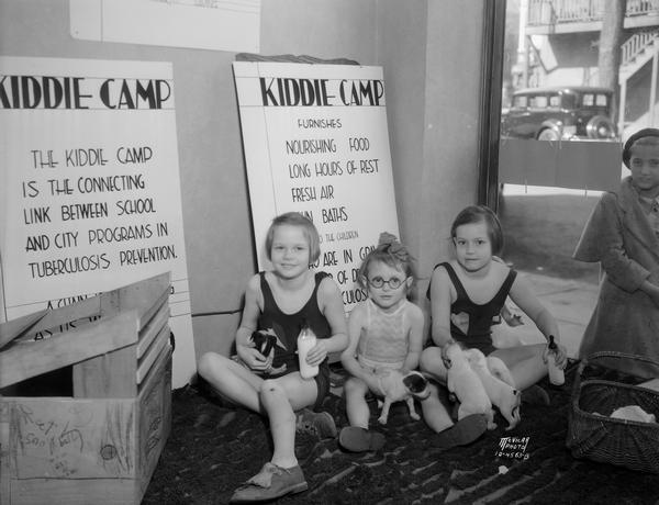 Three little girls, members of the Emerson Fresh Air school and the Longfellow Nutrition room, playing with puppies in the <i>Capital Times</i> Kiddie Camp display window at 116 N. Fairchild Street. They are there to demonstrate what the Kiddie Camp will mean to "pre-tubercular" children.