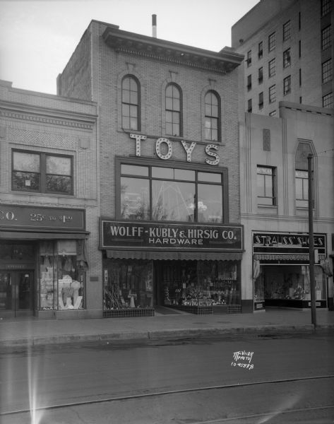 Wolff-Kubly & Hirsig Co. hardware store, located at 17 South Pinckney Street. There is a lighted "TOYS" sign above the second floor display window. The view also includes Strauss' Shoes on the right, located at 19 South Pinckney Street.