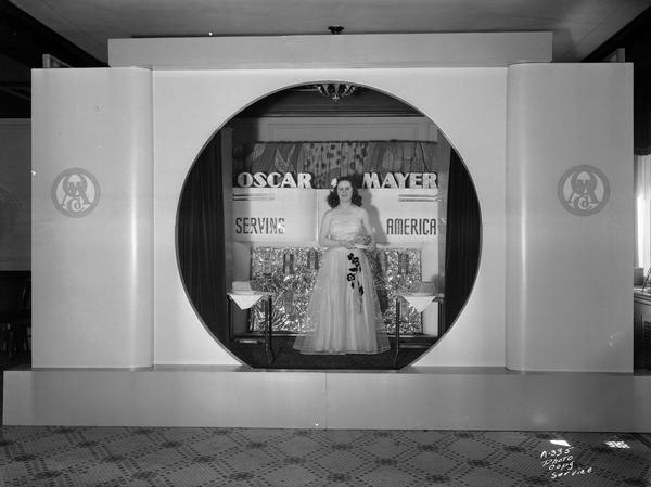 A female model standing and wearing a long dress on the "Oscar Mayer Serving America" stage set, holding a meat product.