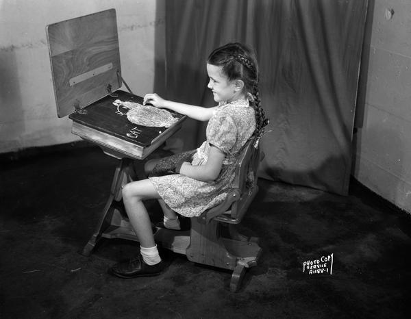 Young girl seated at child's training desk with top open showing blackboard.