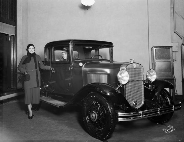 View of a girl from an Orpheum act posing with new Chevrolet coupe parked indoors. There is a man sitting in the passenger seat.