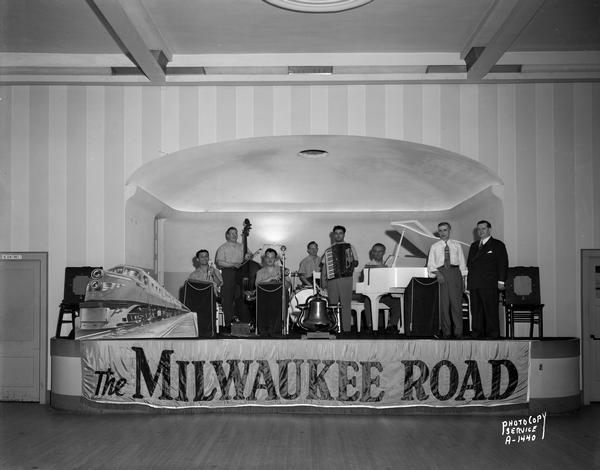 Milwaukee Road Service Club Orchestra performing on stage at American Legion Memorial Hall, 110 East Wilson Street. Decorations include large bell, Milwaukee Road engine and large sign: "The Milwaukee Road."