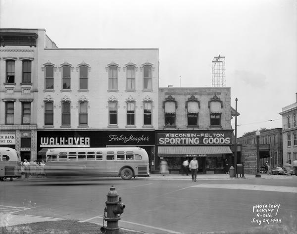Businesses along West Main Street during the remodeling of the Blied Building, 29 West Main Street, after the top one and two stories have been removed. Businesses shown are Madison Bank and Trust, Walk-Over Shoe Store, Forbes-Meagher Music Store, and Wisconsin-Felton Sporting Goods. Also in the photograph are pedestrians and buses.