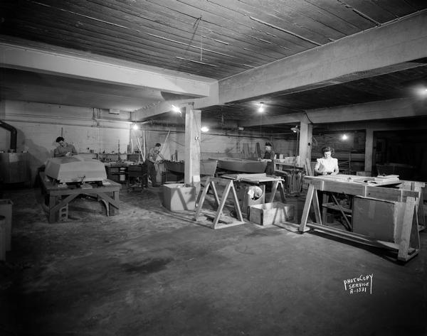 Inland Lakes Boat Works basement workshop at the Stadium Garage, located at 1501 Monroe Street. The view includes three men and one woman constructing boats.
