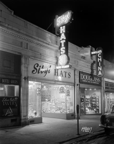 Night view of the "Mifflin Arcade" with terra cotta facade. Businesses include Silver Dollar Tavern at 117 West Mifflin Street, Sibyl Hats at 119 West Mifflin Street, Douglas China and Glassware at 121 West Mifflin Street, and Home Makers Appliance at 123 West Mifflin Street. Neon signs for Sibyl and Douglas are lit up.  Sign for Sibyl Hat Shop says "Hats, $1.99, $2.99, None Higher."
