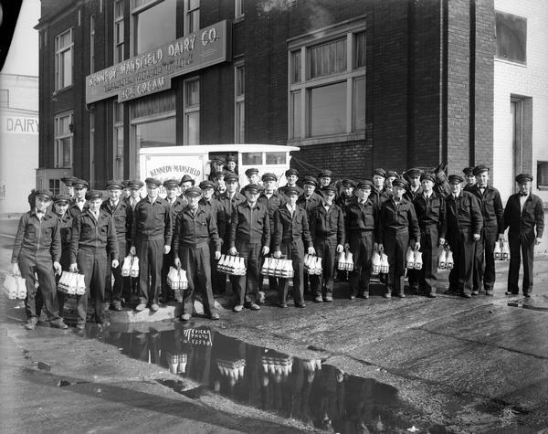 Group portrait of Kennedy Mansfield milk men in uniform carrying milk bottles. They are standing in front of a horse-drawn milk wagon and the Kennedy Mansfield Dairy building, located at 621-629 West Washington Avenue.