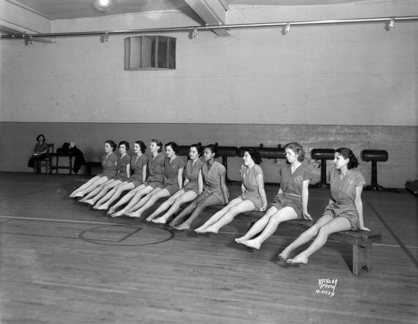 Ten Central High School girls in gym suits sit on benches doing foot exercises.