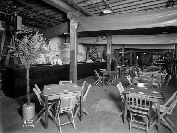 Palm Garden refreshment stand with three men behind a bar, with tables and chairs, and decorative palm trees. The stand was set up at the East Side Business Men's Association Fall Festival, Fuller & Johnson Building, located at 1402 East Washington Avenue.