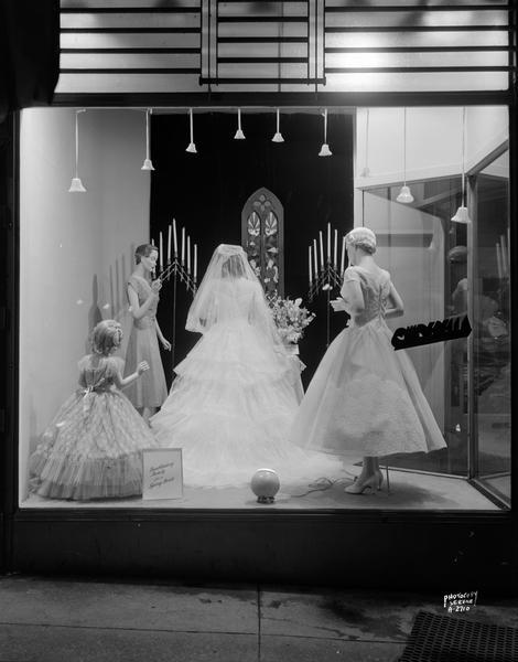 Brides window at the Cinderella Frocks store, located at 8 South Carroll Street. In the window are four mannequins dressed in bridal attire in an altar scene with window, candelabra, and vase of flowers.