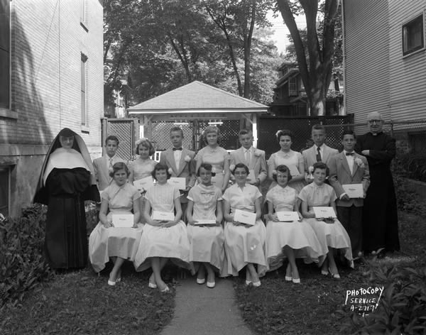 Outdoor group portrait of a graduation class of 9 girls and 5 boys, holding their diplomas, with a priest and a nun at Holy Redeemer Church, located at 128 West Johnson Street.