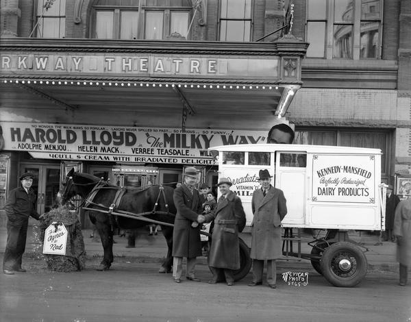 View from street of Barney Miller, the driver, feeding hay to Ned, the oldest dairy horse in service in Madison. The horse is hitched to a Kennedy Dairy wagon in front of Parkway Theater, 6-10 W, Mifflin Street. E.A. Babcock and H.H. Mellinger of Kennedy-Mansfield Co., acknowledge Ned's gift of hay by shaking hands with John Scharnberg, manager of the Parkway. The marquee advertises Harold Lloyd in "The Milky Way."