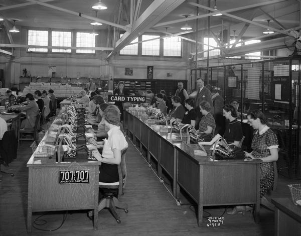 Wisconsin Auto License Bureau Card Typing Department, located at 16-20 East Doty Street. Women government employees are sitting at desks, while a male supervisor and Secretary of State Fred Zimmerman, the government official then responsible for issuing Wisconsin drivers licenses, stand in the background. The state of Wisconsin issued its first drivers license in 1905. The only decoration in this sterile workplace is a license plate with the slogan "America's Dairyland."