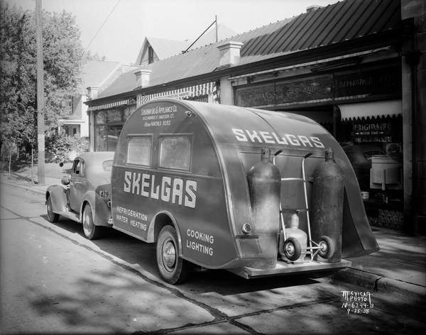 Three-quarter view from left rear of a Skelgas trailer, showing two gas tanks, being pulled by an automobile, parked in front of Suburban Gas & Appliance Co., 1923 Monroe Street.