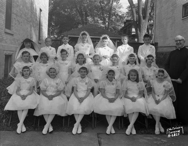 Outdoor group portrait of first communion class of Holy Redeemer Catholic Church, 128 West Johnson Street. The group is near the church and includes sixteen girls, four boys, one nun and one priest.