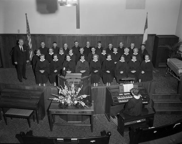 Elevated group portrait of Bethany Lutheran Church choir in choir robes taken inside the church, 301 Riverside Drive. A woman sits on a bench in front of the church organ in the foreground.
