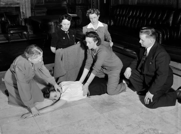 Artificial respiration being taught in a Red Cross first aid class to State employees in the Wisconsin State Capitol Assembly Parlor, supervised by Arne Lerwick (far right), instructor from the Madison Fire Department. Mrs. Ralph Brown, State Beverage Tax Department employee, administers treatment to the victim, Margaret Redmond, Beverage Tax Dept., with the help of Marion Poole, instructor, Highway Commission.  Looking on are Margarite Bernardy, Motor Vehicle Department, and Esther Schadde, instructor Board of Health.